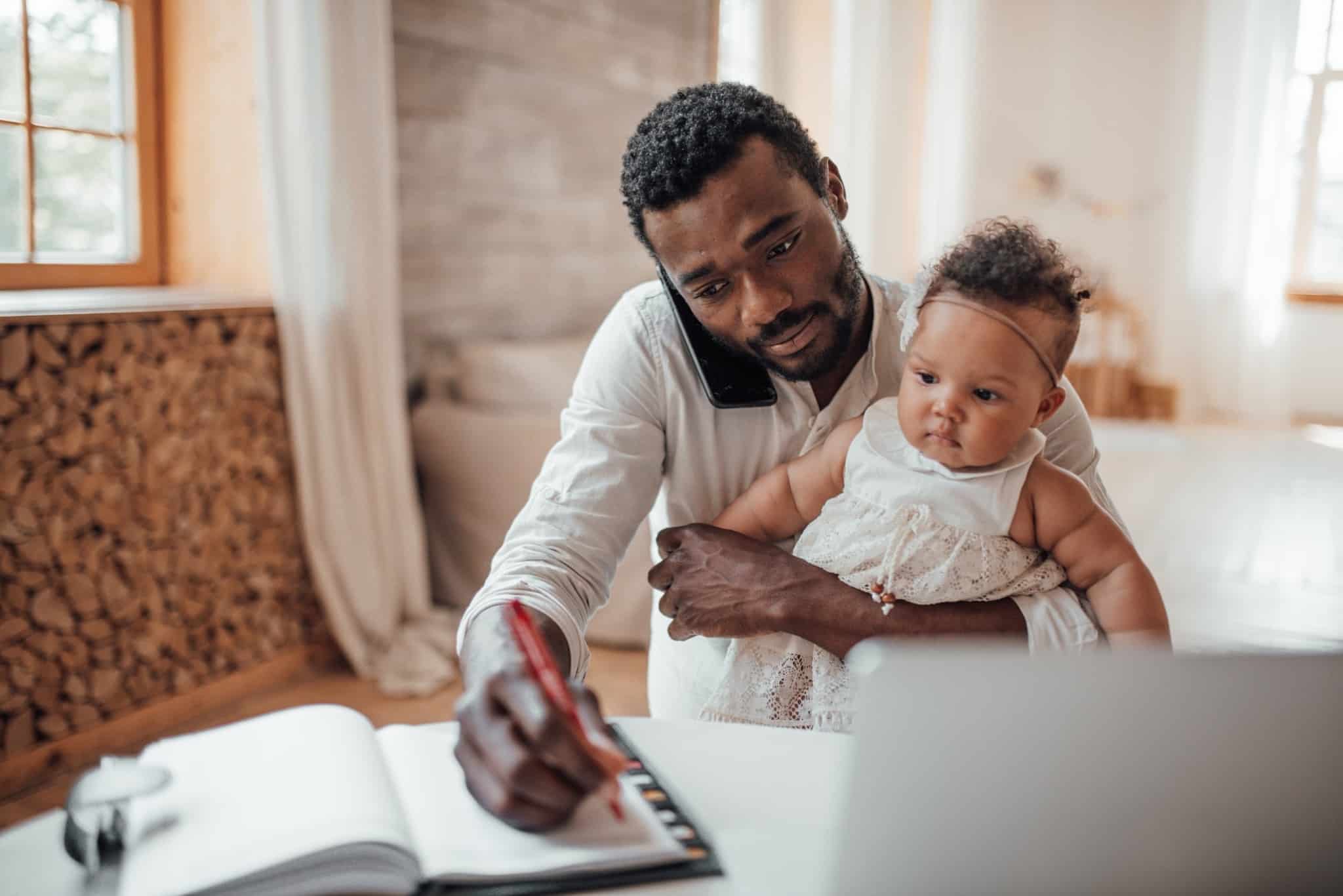Image of a father with his little daughter on his lap, at the table, with a laptop, while writing in a notebook - an allusion to the IRS for divorced parents