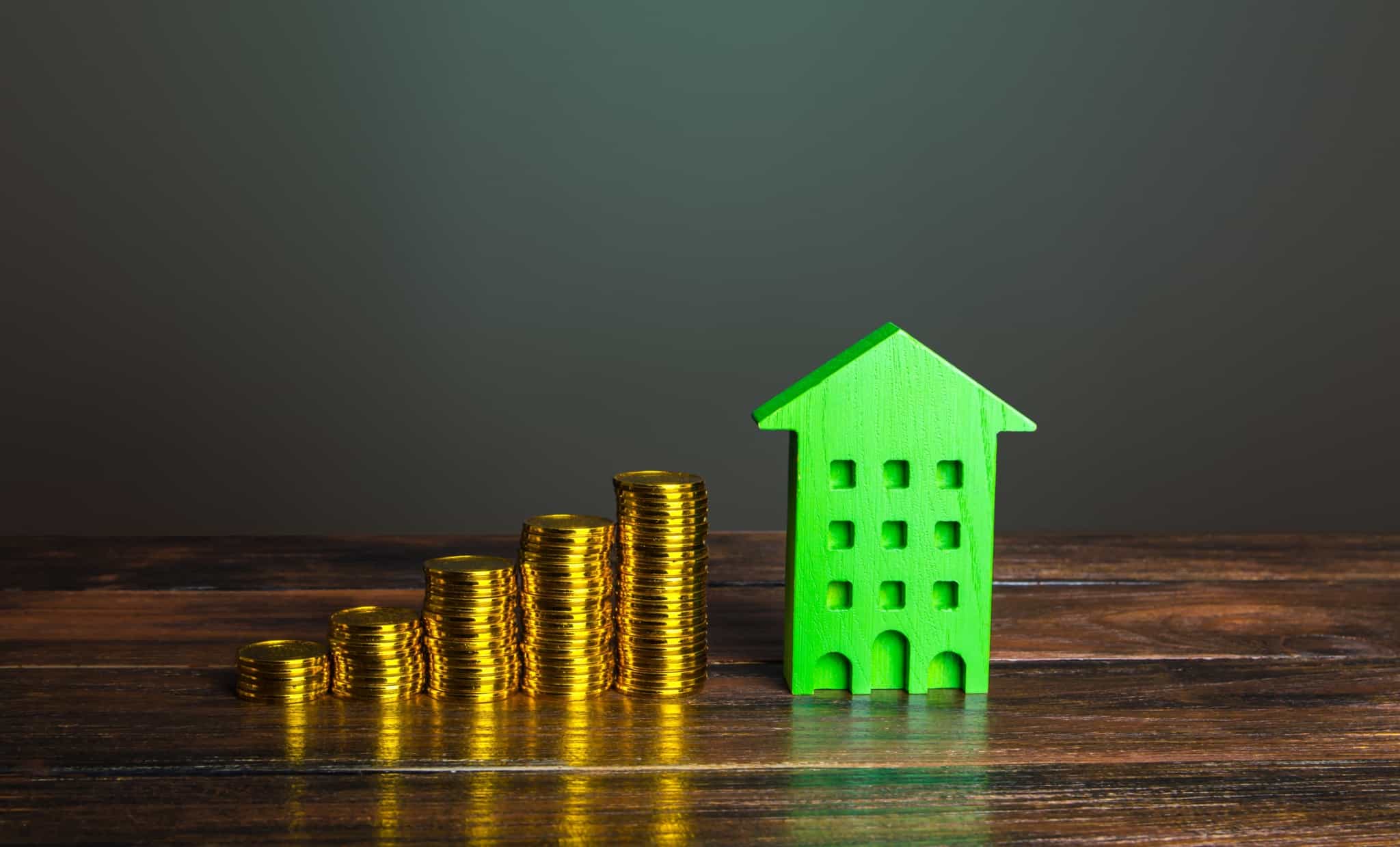 photographic production shows, on a dark background, a model of the facade of a building, in wood and green to point to sustainability, and beside it growing piles of coins, pointing to savings.  the set illustrates the need to invest in energy efficiency